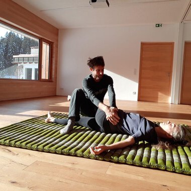 Eine Person liegt auf einer Matte, während eine andere Person sie sanft behandelt. Der Raum hat große Fenster mit Blick auf Bäume und viel Tageslicht. | © Alexander Mayr