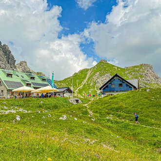 Eine malerische Berglandschaft mit gemütlichen Hütten und grünen Wiesen. Im Hintergrund sind beeindruckende Felsen und ein blauer Himmel mit Wolken zu sehen. | © Mindelheimer Hütte | Wolfgang Stache