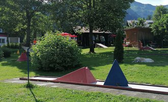 Ein Minigolfplatz mit bunten Hindernissen in einer grünen Umgebung. Im Hintergrund sind Bäume und ein Gebäudeteil zu sehen.
