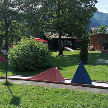 Ein Minigolfplatz mit bunten Hindernissen in einer grünen Umgebung. Im Hintergrund sind Bäume und ein Gebäudeteil zu sehen.