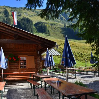 © Kleinwalsertal Tourismus Eine gemütliche Hütte in den Bergen mit Tischen und Bänken im Freien. Im Hintergrund sind grüne Wiesen und Berge zu sehen. | © Kleinwalsertal Tourismus