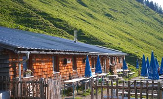 Eine rustikale Almhütte mit Holzverkleidung und einem schrägen Dach. Im Vordergrund gibt es Tische mit blauen Sonnenschirmen in einer grünen Wiesenlandschaft. | © Kleinwalsertal Tourismus