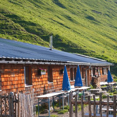 Eine rustikale Almhütte mit Holzverkleidung und einem schrägen Dach. Im Vordergrund gibt es Tische mit blauen Sonnenschirmen in einer grünen Wiesenlandschaft. | © Kleinwalsertal Tourismus