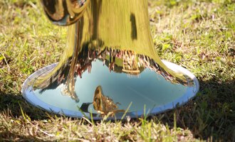 Eine glänzende Trompete, die auf dem Gras steht. Im reflektierenden Metall sind Bäume und Menschen zu sehen. | © Kleinwalsertal Tourismus | Frank Drechsel