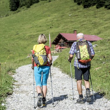 A couple is walking on a gravel path in a green landscape. In the background, a small hut can be seen. | © Kleinwalsertal Tourismus | Dominik Berchtold