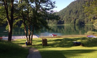 Ein ruhiger See umgeben von Bäumen und grünen Wiesen. Im Hintergrund sind Berge und Sonnenliegen am Ufer zu sehen. | © Kleinwalsertal Tourismus | Naturbad Freibergsee