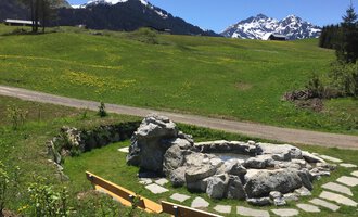 A quiet, green landscape area with a stone bench and mountain view. The sun is shining on a clear day. | © Kleinwalsertal Tourismus | Sarina Berchtold