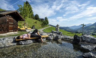 Eine entspannende Szene in den Bergen mit einer Person, die am Ufer eines kleinen Teiches liegt. Im Hintergrund sind grüne Hügel und ein klarer Himmel zu sehen. | © Kleinwalsertal Tourismus | Dominik Berchtold