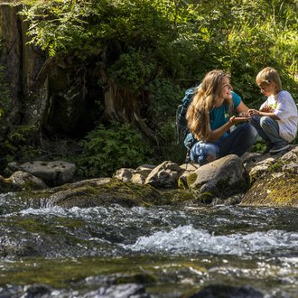 Eine Mutter und ihr Sohn sitzen an einem klaren Bach in der Natur. Sie haben Spaß und genießen die Zeit miteinander. | © Kleinwalsertal Tourismus | Frank Drechsel
