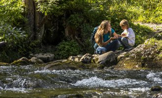 Eine Mutter und ihr Sohn sitzen an einem klaren Bach in der Natur. Sie haben Spaß und genießen die Zeit miteinander. | © Kleinwalsertal Tourismus | Frank Drechsel