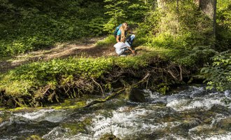 Zwei Personen sitzen am Ufer eines plätschernden Bachs, umgeben von grüner Natur. Die Sonne scheint durch die Bäume, und es herrscht eine ruhige Atmosphäre. | © Kleinwalsertal Tourismus | Frank Drechsel