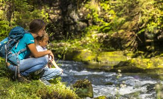 Eine Frau sitzt mit einem Kind am Ufer eines plätschernden Bachs. Beide genießen die Natur und die ruhige Umgebung. | © Kleinwalsertal Tourismus | Frank Drechsel