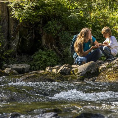 Eine Mutter und ihr Sohn sitzen an einem klaren Bach in der Natur. Sie haben Spaß und genießen die Zeit miteinander. | © Kleinwalsertal Tourismus | Frank Drechsel