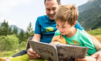 Ein Vater und sein Sohn sitzen im Freien und lesen gemeinsam ein Buch. Beide lächeln und der Junge hält ein Stofftier in der Hand. | © Kleinwalsertal Tourismus | Bastian Morell
