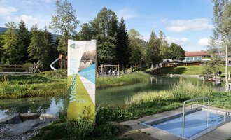 Ein kleiner Swimmingpool in einer grünen Wiese, umgeben von Bäumen. Im Hintergrund sind Spielgeräte und ein Gebäude zu sehen. | © Kleinwalsertal Tourismus | Veronika Senn