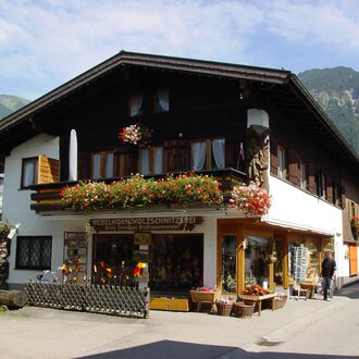 © Nebelhorn Holzschnitzerei e.K. A traditional house in the mountains, surrounded by greenery. The shop on the ground floor offers handcrafted wooden carvings and is adorned with flowers. | © Nebelhorn Holzschnitzerei e.K.