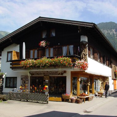 A traditional house in the mountains, surrounded by greenery. The shop on the ground floor offers handcrafted wooden carvings and is adorned with flowers. | © Nebelhorn Holzschnitzerei e.K.