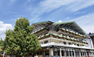 Ein charmantes Hotel mit Balkonen und bunten Pflanzen. Im Vordergrund steht ein großer Baum unter einem klaren blauen Himmel. | © Kleinwalsertal Tourismus | N. Lughammer