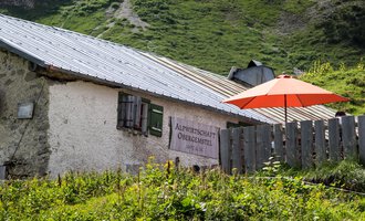 Eine charmante Almhütte mit einem roten Sonnenschirm im Grünen. Im Hintergrund sind sanfte Hügel und eine Berglandschaft zu sehen. | © Kleinwalsertal Tourismus | Andre Tappe