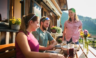 Eine freundliche Atmosphäre im Freien mit zwei Gästen an einem Tisch. Eine Bedienung serviert das Essen in einem hübschen, rustikalen Lokal. | © Kleinwalsertal Tourismus | Oliver Farys