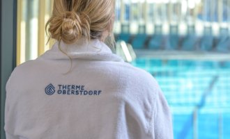 A person in a white bathrobe with the logo "Therme Oberstdorf" stands in front of a swimming pool. A relaxing atmosphere is visible. | © Tourismus Oberstdorf
