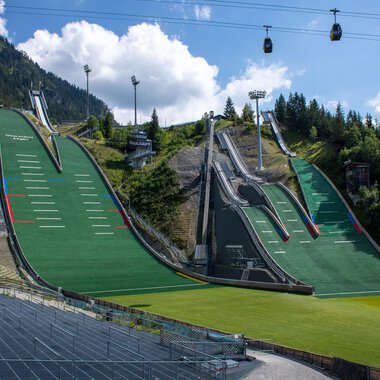 Two large ski jumps surrounded by green meadows and mountains. In the background, cable cars hang under a blue sky. | © Skiclub Oberstdorf Veranstaltungs GmbH