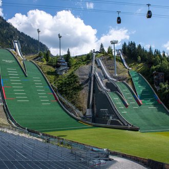 Two large ski jumps surrounded by green meadows and mountains. In the background, cable cars hang under a blue sky. | © Skiclub Oberstdorf Veranstaltungs GmbH