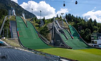 Two large ski jumps surrounded by green meadows and mountains. In the background, cable cars hang under a blue sky. | © Skiclub Oberstdorf Veranstaltungs GmbH