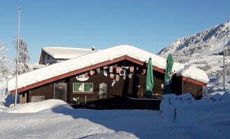 A cozy wooden house with a snow-covered roof in a wintry landscape. In the background, there are snow-covered mountains and a blue sky. | © Panoramahütte