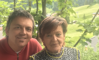 Ein Mann und eine Frau stehen zusammen in der Natur. Im Hintergrund sind grüne Hügel und Bäume zu sehen.