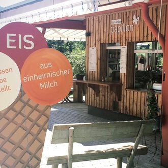 Eine gemütliche Eisdiele mit Holzverkleidung und einem Thekenbereich. Im Vordergrund steht ein großes Eisschild, das auf hausgemachtes Eis hinweist. | © PAPILLON EISDIELE | Eis & Crêpe | Lisa Schüttler