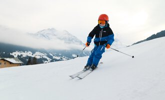Ein Junge fährt Ski auf einer verschneiten Piste. Im Hintergrund sind Berge und Wolken zu sehen. | © Kleinwalsertal Tourismus | Oliver Farys