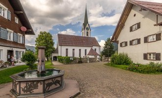 A picturesque village square with a fountain in the foreground and traditional houses. In the background, there is a church with a tall tower and a cloudy sky. | © Kleinwalsertal Tourismus | Steffen Berschin