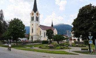 A picturesque church with a tall tower stands next to a charming building. The sky is partly cloudy and the surroundings are green and inviting. | © Kleinwalsertal Tourismus | Oliver Farys