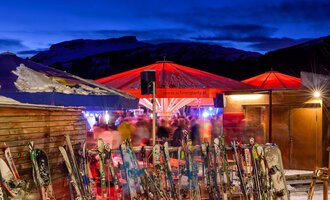 A ski hut at night with red-lit parasols and many skis in the foreground. In the background, blurred people can be seen enjoying a convivial atmosphere. | © Plattform Kleinwalsertal | Michael Monschau