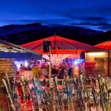 A ski hut at night with red-lit parasols and many skis in the foreground. In the background, blurred people can be seen enjoying a convivial atmosphere. | © Plattform Kleinwalsertal | Michael Monschau