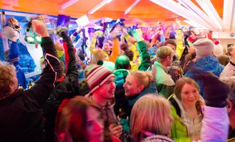 A lively celebration with many people in colorful ski outfits. The atmosphere is energetic with lights and cheerful dancing. | © Plattform Kleinwalsertal | Michael Monschau