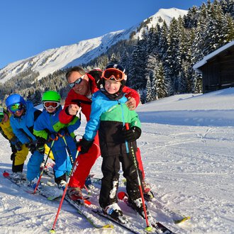 Eine Gruppe von Kindern und einem Erwachsenen beim Skifahren in der verschneiten Berglandschaft. Sie tragen bunte Skikleidung und lächeln in die Kamera. | © Andy Herr | Werbewind GmbH