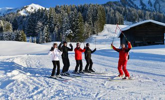 Eine Gruppe von fünf Skifahrern steht fröhlich im Schnee und genießt den sonnigen Tag. Im Hintergrund sind schneebedeckte Bäume und ein Holzhaus zu sehen. | © Andy Herr | Werbewind GmbH