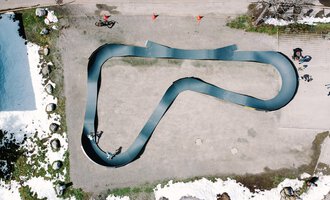 A top view of a curved racetrack made of black material. Around the track, some people and remnants of snow can be seen. | © Christian Posch | Klaus Zopf