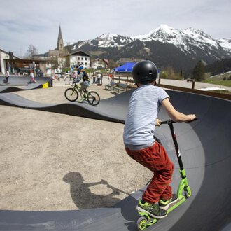© Kleinwalsertal Tourismus | Frank Drechsel Ein Skatepark mit Kindern, die auf einem Roller und Fahrrad fahren. Im Hintergrund sind Berge und eine kleine Stadt zu sehen. | © Kleinwalsertal Tourismus | Frank Drechsel