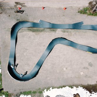 A top view of a curved racetrack made of black material. Around the track, some people and remnants of snow can be seen. | © Christian Posch | Klaus Zopf