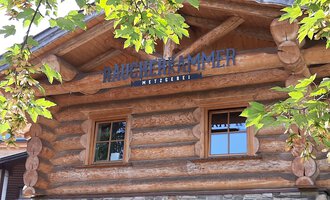 A rustic wooden building with a sign that reads "Mühlenkammer." Surrounded by green leaves and a clear sky. | © Kleinwalsertal Tourismus | Veronika Senn