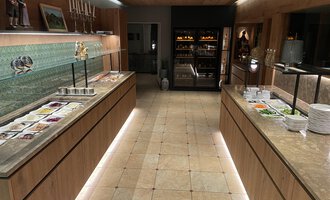 Ein modern gestalteter Buffetbereich mit Holzregalen und einer schönen Glasfront. Auf den Tischen sind verschiedene Speisen und frische Zutaten angerichtet. | © Almhof Rupp | Lukas Kind