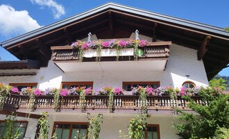 Ein traditionelles Alpenhaus mit blühenden Balkonpflanzen. Der Himmel ist blau und die Umgebung ist grün. | © Kleinwalsertal Tourismus | N. Lughammer