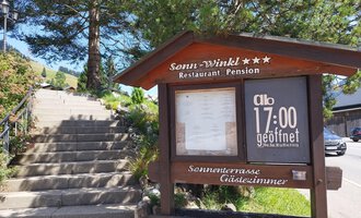 Ein Holzschild des Restaurants "Sonu-Winkl" zeigt die Öffnungszeiten an. Im Hintergrund sind eine Treppe und Bäume zu sehen. | © Kleinwalsertal Tourismus | N. Lughammer