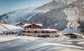 Ein malerisches Bergdorf im Winter mit schneebedeckten Hütten. Die Umgebung zeigt hohe, verschneite Berge und einen klaren Himmel.