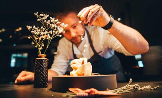 A cook decorates a dish with a lot of love and skill. In the background, there is a small flower in a vase. | © Hotel Birkenhöhe | Stefan Klauser