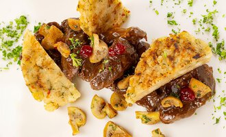 Ein köstliches Gericht mit Fleischstücken in einer dunklen Soße, garniert mit Pilzen und Preiselbeeren. Dazu sind knusprige Beilagen angerichtet. | © Hotel Alte Krone