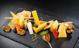 Eine Käseplatte mit verschiedenen Käsesorten, Nüssen und frischen Früchten. Dazu gibt es einen Löffel mit einer fruchtigen Marmelade auf einem schwarzen Teller. | © Hotel Alte Krone